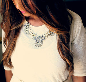 Image of beautiful brunette with long brown hair with chestnut highlights, illustrating what could be possible using LUSHIERE clip in hair extensions. Model is looking down and wearing an ornate necklace, white top and pink lipstick. 
